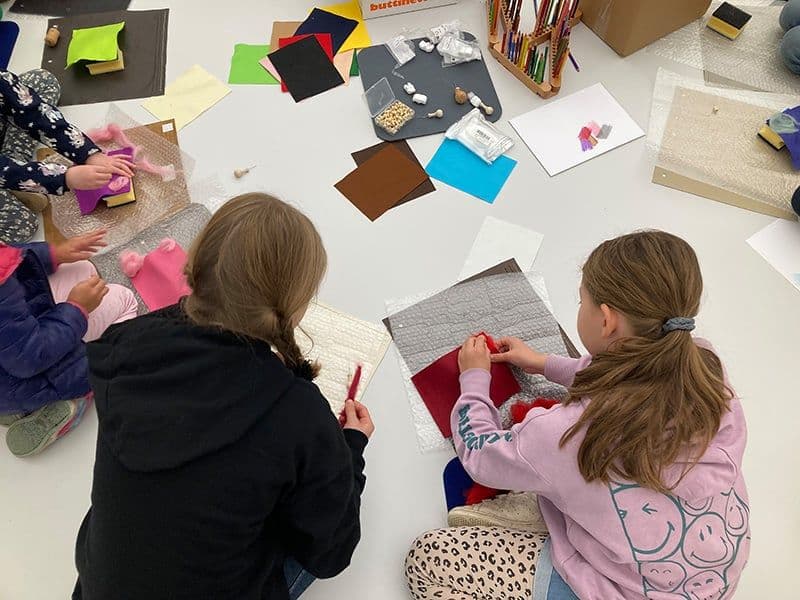 Auf dem Boden sitzen zwei Kinder. Die Kinder basteln mit verschiedenen Materialien.