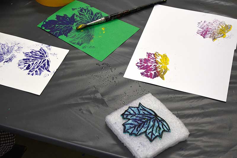 Ein gebastelter Stempel liegt auf einem Tisch. Der Stempel sieht aus wie ein Blatt von einem Baum. Weiße Blätter sind mit dem Stempel bedruckt.