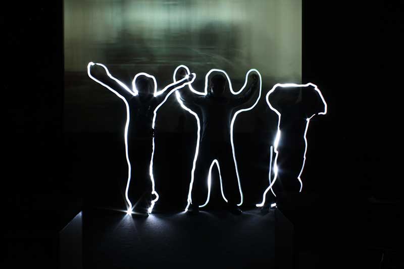 Zu sehen sind drei Formen aus Licht. Das Licht zeichnet die Umrisse von Kindern nach.