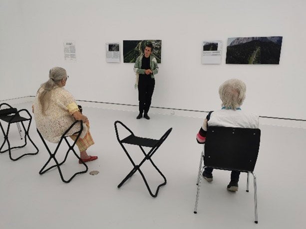 Eine Person steht vor einem Bild. Die Person erzählt etwas zu dem Bild. Zwei ältere Menschen hören ihr zu. Die älteren Menschen sitzen auf Stühlen.