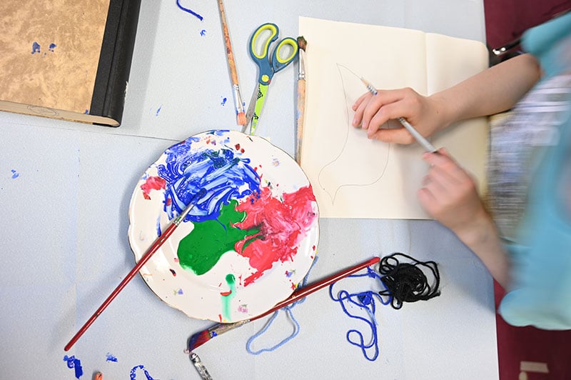 Auf einem Tisch steht ein Teller mit bunten Farben. Neben den Farben liegen Pinsel. Ein Kind malt etwas.
