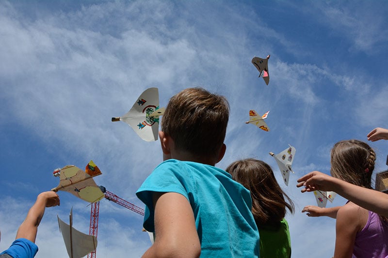 Eine Gruppe von Kindern schaut in den blauen Himmel. Die Kinder werfen Papierflieger in die Luft. Die Papierflieger sind bemalt.